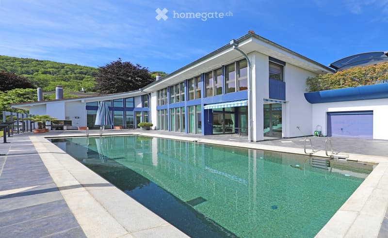 Modern two-story house with large windows, a swimming pool, and a garage. The house features a sleek design with white and blue accents, surrounded by greenery and a clear blue sky.