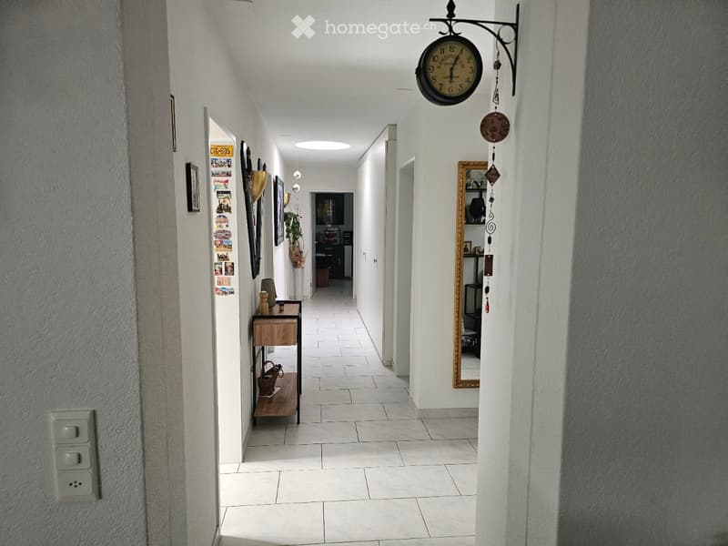 A long, well-lit hallway with white walls, tiled flooring, and several doors leading to different rooms. A small wooden table with a lamp and decorative items is placed on the left side, and a wall-mounted clock with a decorative wind chime hangs on the r
