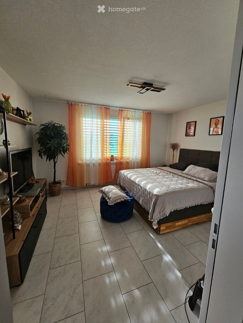 A well-lit bedroom with a large bed, nightstand, ottoman, television, plant, and shelves. The room features tiled flooring and large windows with orange curtains.