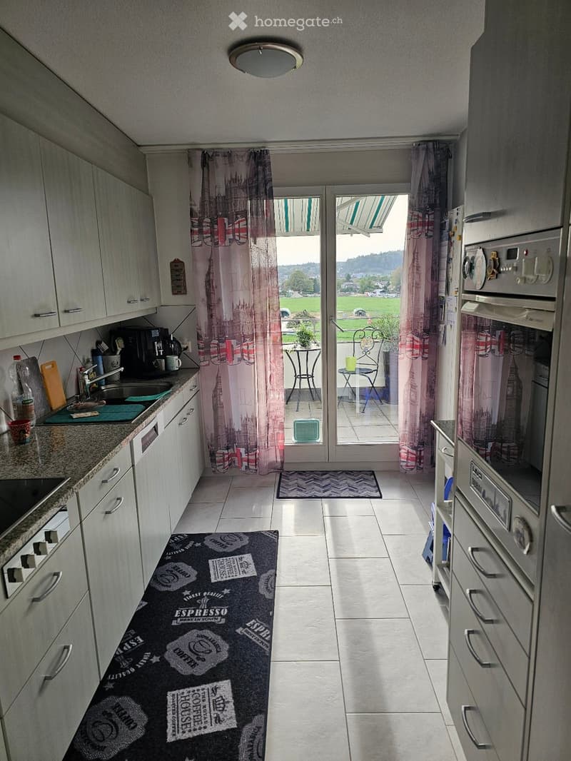 Modern kitchen with white cabinets, granite countertops, and a black rug with coffee-themed design. Features a sink, oven, refrigerator, microwave, and a small table and chairs on a balcony.