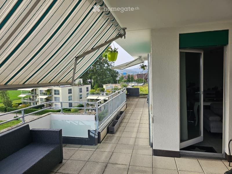 Balcony with a retractable awning, tiled floor, metal railing, and a view of the surrounding area. A wicker sofa is placed on the balcony, and there is a hanging plant.