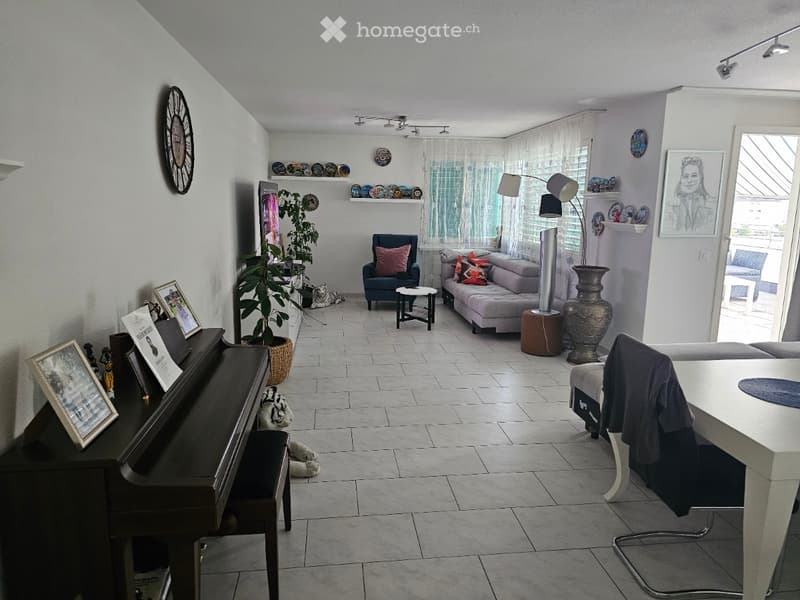 Bright living room with tiled floor, large windows, sofa, armchair, coffee table, piano, floor lamp, plant, and decorative items on shelves.
