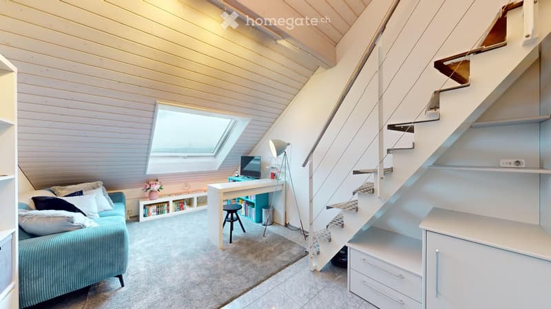 Bright, modern attic room with a slanted wooden ceiling, skylight window, and built-in staircase leading to another level. Features a blue sofa, white desk with laptop, bookshelf, and floor lamp.