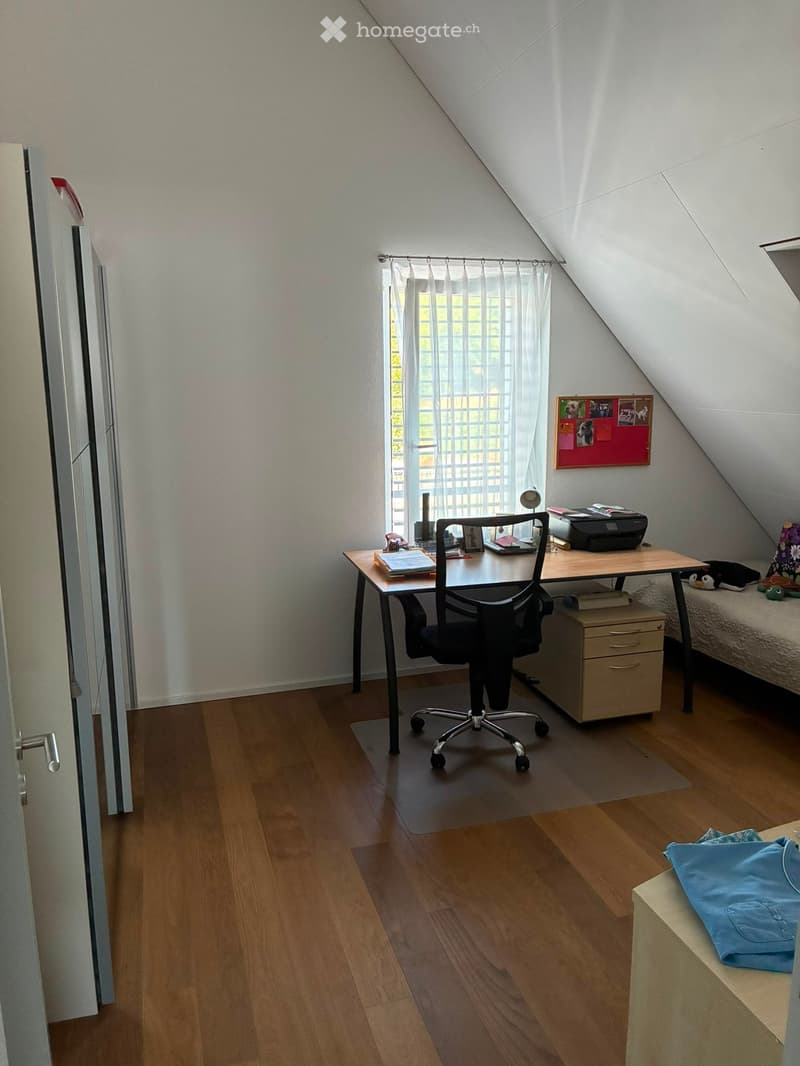 A cozy attic room with wooden flooring, a desk with a chair, a filing cabinet, a small bed with pillows, and a window with white curtains. The room also features a bulletin board, a lamp, and a small stool with a blue cloth on it.