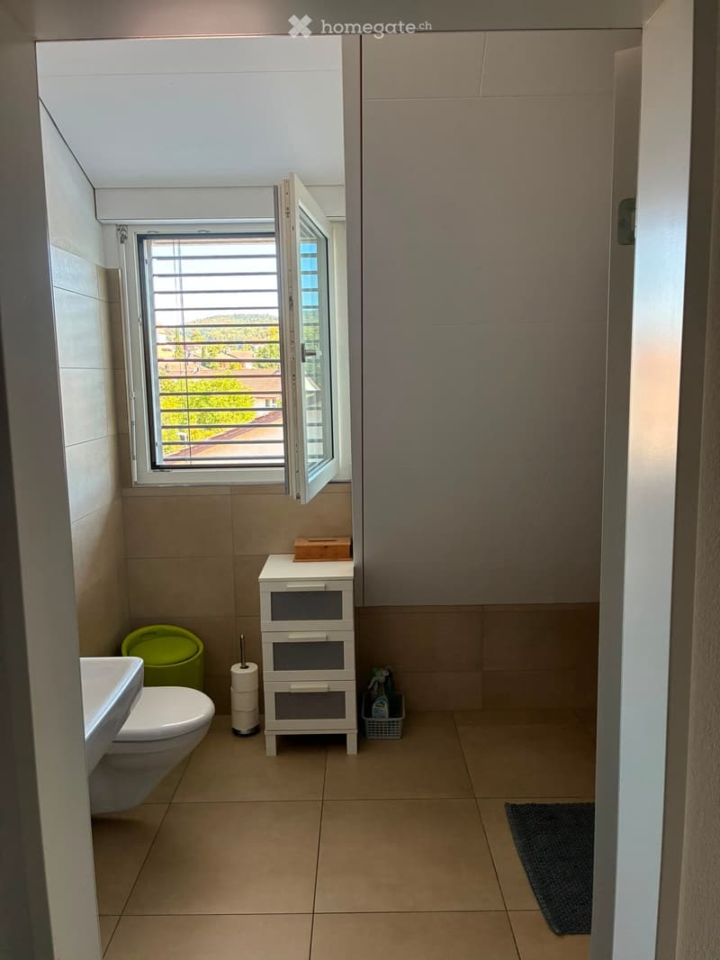 Modern bathroom with beige tiles, a toilet, sink, small storage cabinet, and a window offering a view of the balcony and surrounding area.