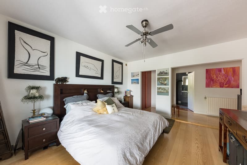 A well-furnished bedroom with a large wooden bed, nightstands, a ceiling fan, and several picture frames on the walls. The room features light wooden flooring and has an open doorway leading to another room.