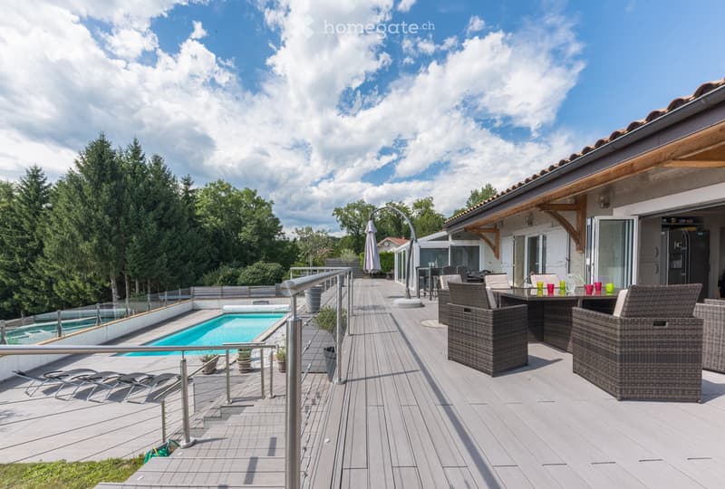 Outdoor patio with a swimming pool, lounge chairs, and a dining area with wicker furniture and colorful drinkware. The area is surrounded by a wooden deck and features a modern glass railing.
