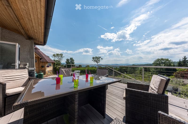 Balcony with a glass table, wicker chairs, and colorful cups, overlooking a scenic landscape with trees and mountains.