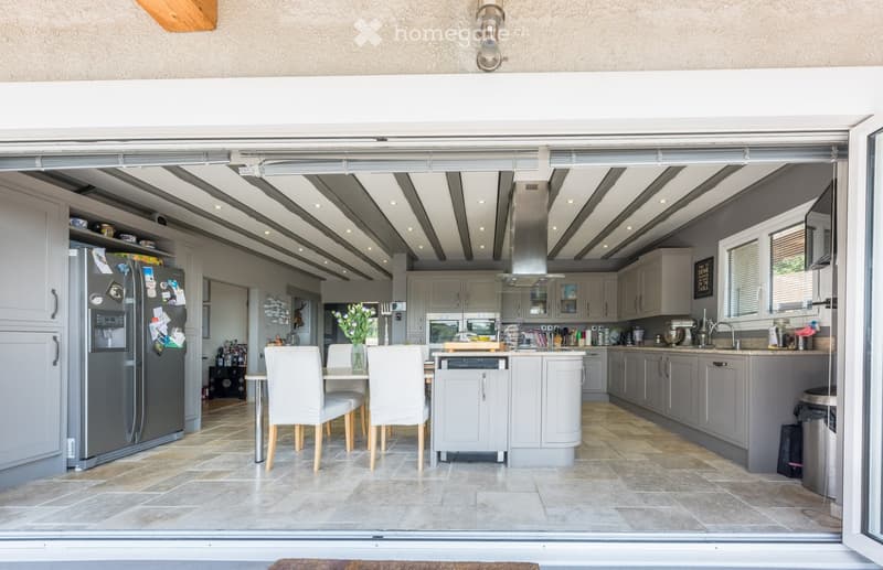 Modern kitchen with grey cabinets, stainless steel appliances, and a central island. Features a dining area with white chairs and a table, and a refrigerator with various magnets and notes.