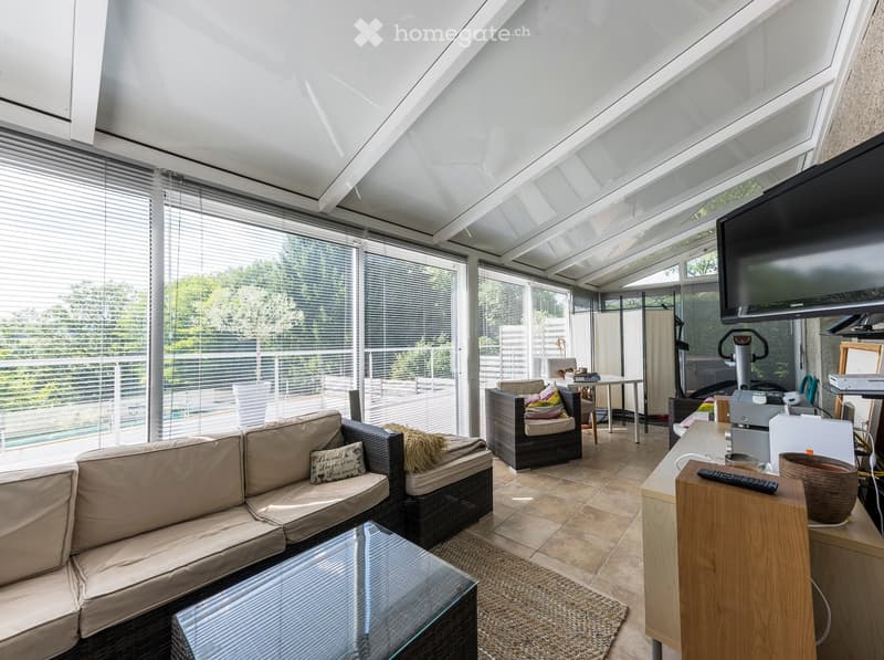 Bright living room with large windows and a view of greenery, featuring a beige sofa, glass coffee table, wicker chairs, and a small table with a laptop and cups.