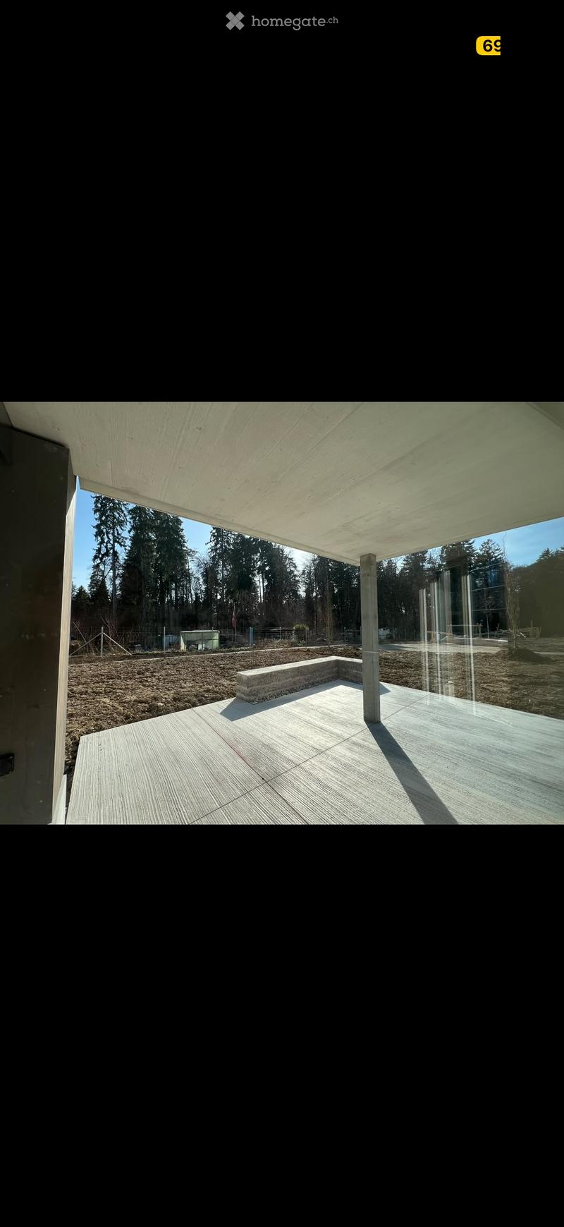 Modern outdoor patio with wooden decking, concrete bench, and large overhanging roof supported by a single column. The area is surrounded by a gravel surface and trees.