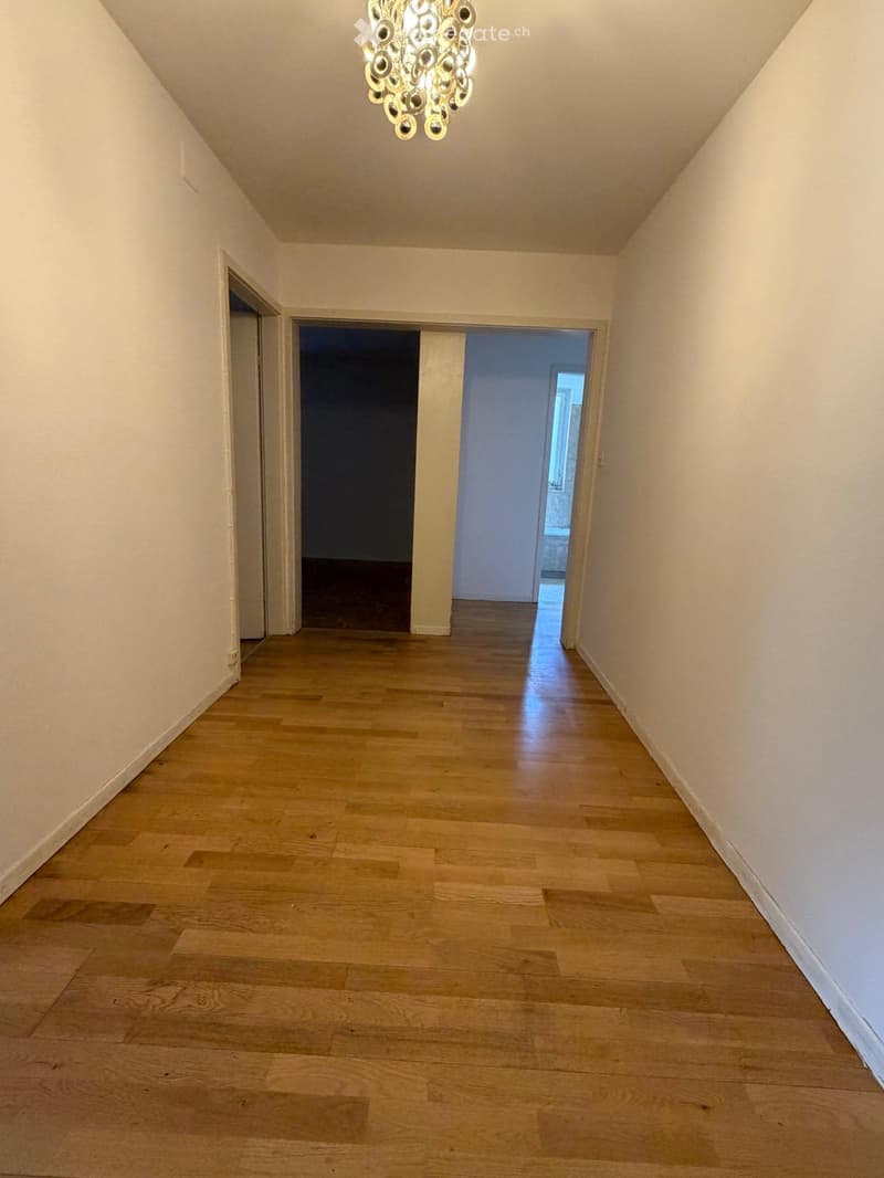 Empty hallway with wooden flooring, white walls, and a decorative chandelier hanging from the ceiling. Two doors lead to other rooms.