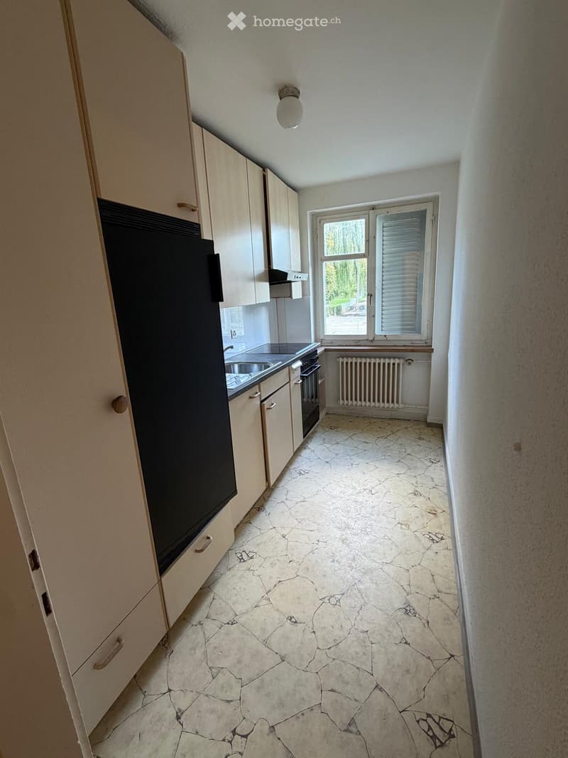 Empty kitchen with light-colored cabinets, a sink, and a window with blinds. The floor is made of cracked stone tiles.