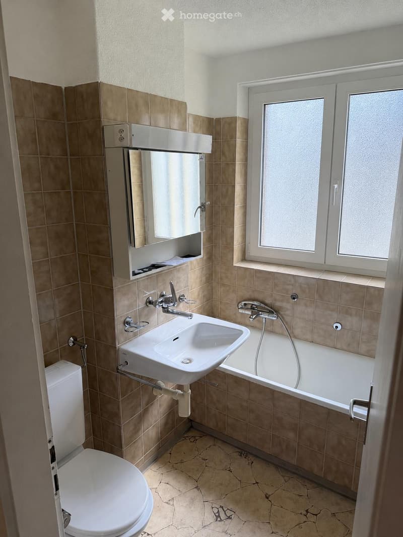 Modern bathroom with beige tiles, a white toilet, a sink with a modern faucet, a bathtub with a handheld shower, and a large mirror above the sink.
