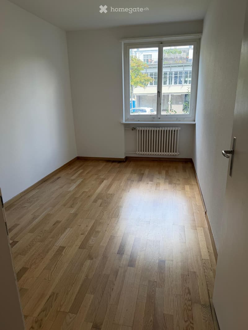 Empty room with wooden flooring, white walls, a window with a radiator below, and a door visible on the right side.