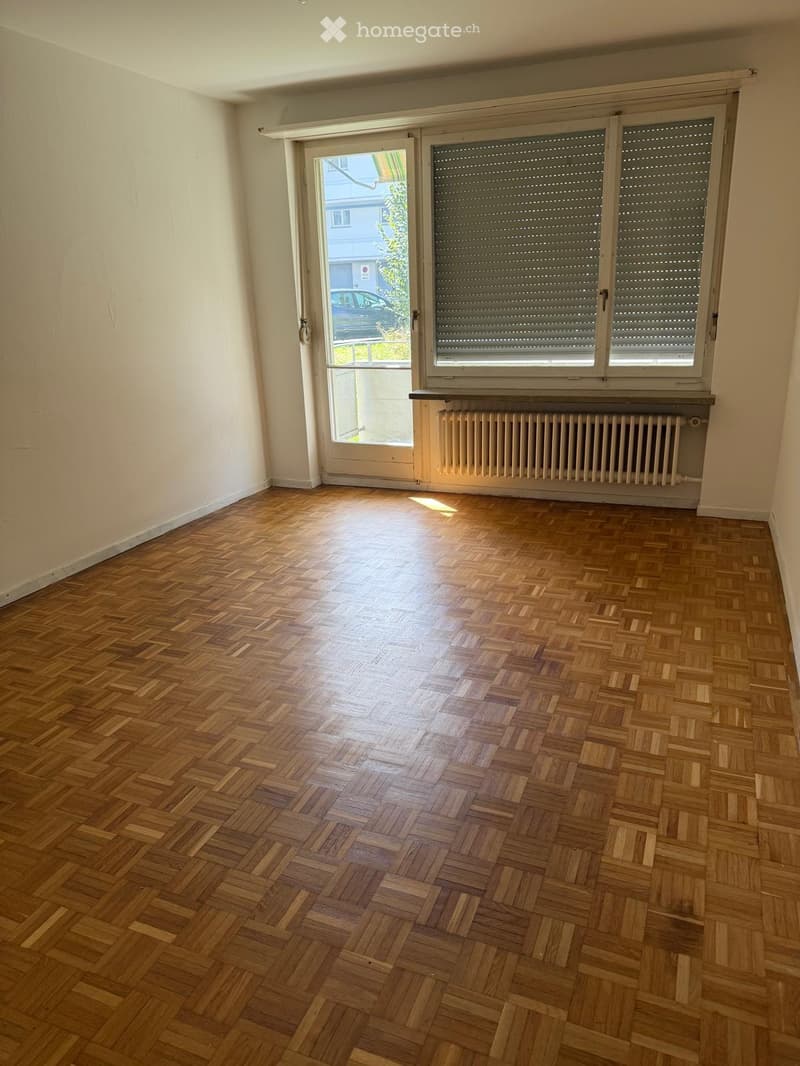 Empty room with wooden herringbone flooring, white walls, and a large window with blinds. A radiator is visible below the window, and a door leads to an outdoor area.