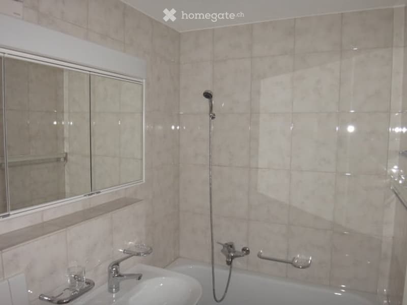 Bathroom with beige tiled walls and floor, a white sink with a silver faucet, a bathtub with a handheld showerhead, a large mirror with a built-in shelf, and a handrail for accessibility.