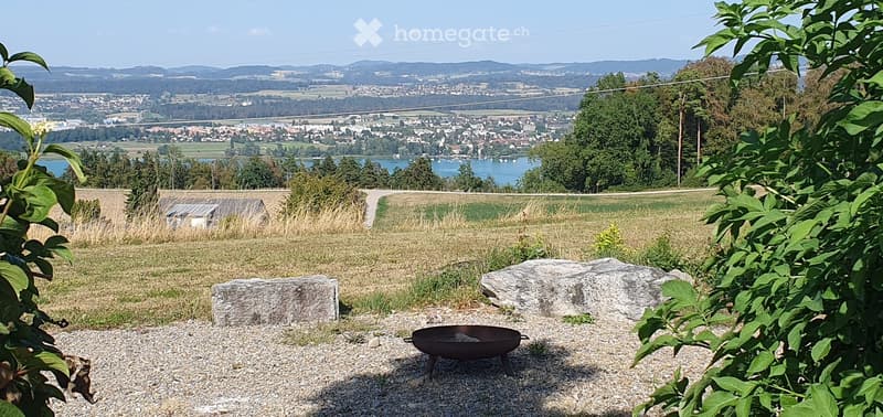 A serene garden area with a fire pit, stone seating, and a panoramic view of a town and lake in the distance.