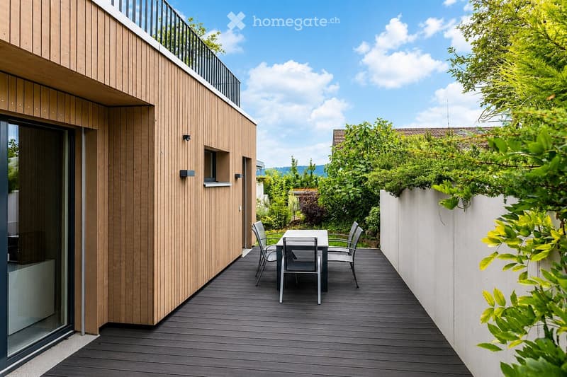 Modern wooden deck terrace with a small table and chairs, surrounded by greenery and a white wall.