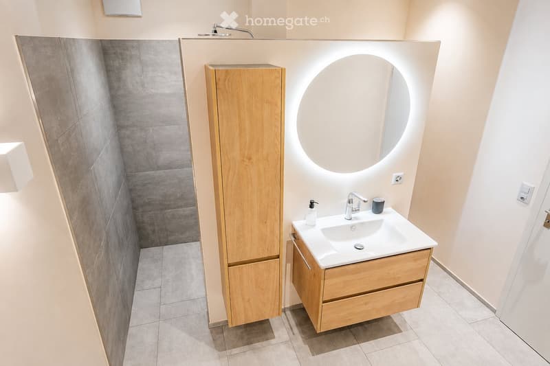 Modern bathroom with a white sink, wooden cabinet, round mirror, and grey tiled walls.