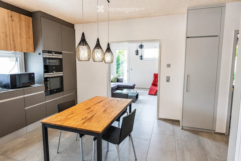 Modern kitchen and dining area with wooden table, chairs, cabinets, refrigerator, oven, microwave, and a living area with a sofa and ottoman.