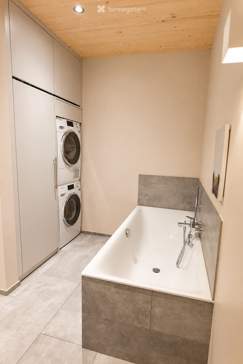 Modern bathroom with a freestanding bathtub, sleek faucet, and a stacked washer/dryer unit. The space features light-colored tiles and a wooden ceiling.