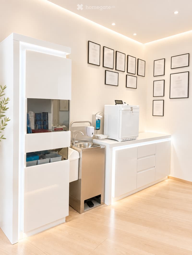 Modern reception area with a white counter, sink, hand sanitizer dispenser, refrigerator, and framed pictures on the wall.