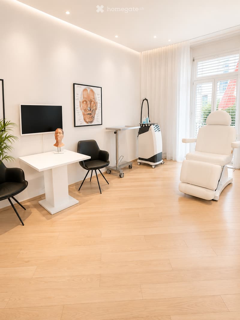 Modern, well-lit room with light wooden flooring, white walls, and large windows with curtains. Features a white table with a mannequin head, two black chairs, a large screen, a medical examination chair, and a small cart with medical equipment.