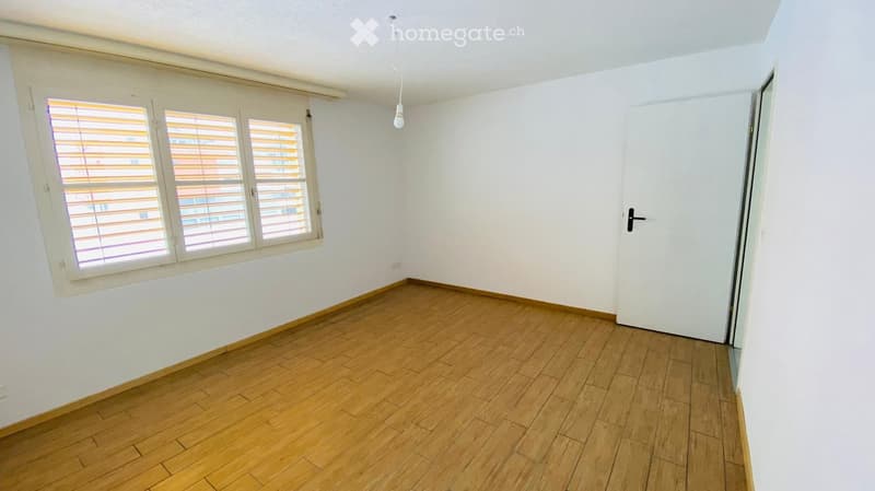 Empty room with wooden flooring, white walls, large window with blinds, and a single door.