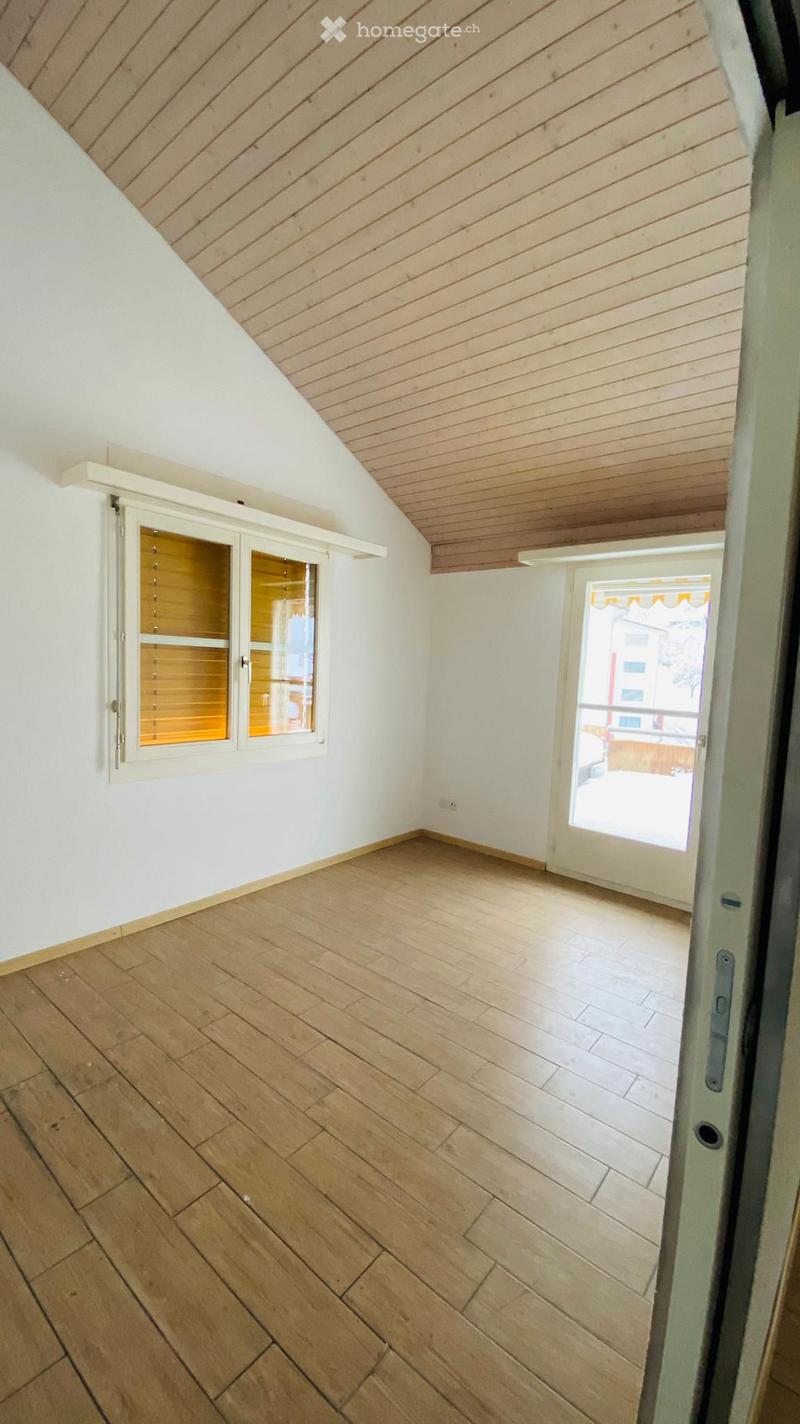Empty room with wooden flooring, white walls, and a slanted wooden ceiling. Features a window with blinds and a sliding glass door leading to a balcony.