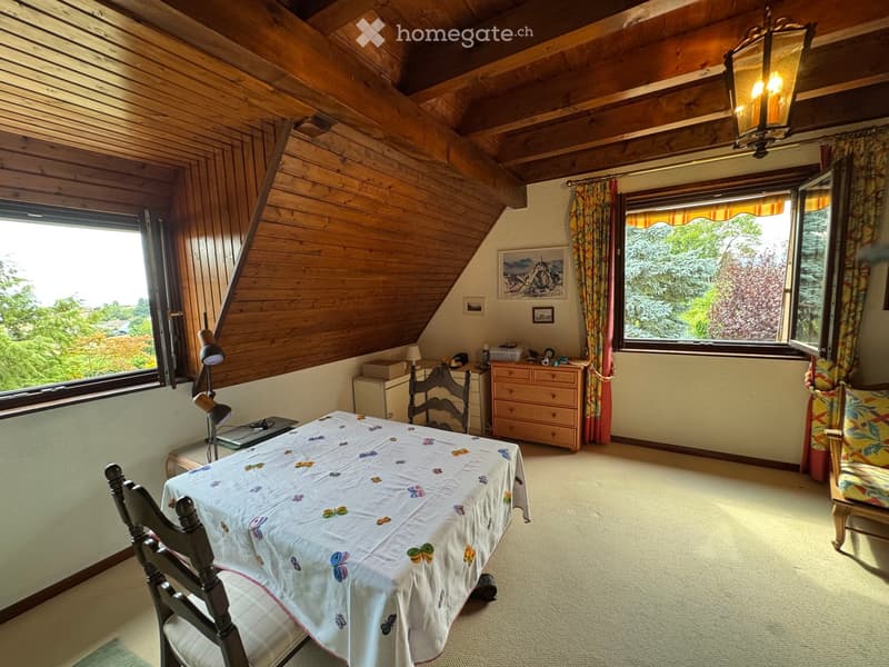 Cozy attic room with wooden beams, large windows, a table with a butterfly-patterned tablecloth, chairs, a lamp, and a chest of drawers.