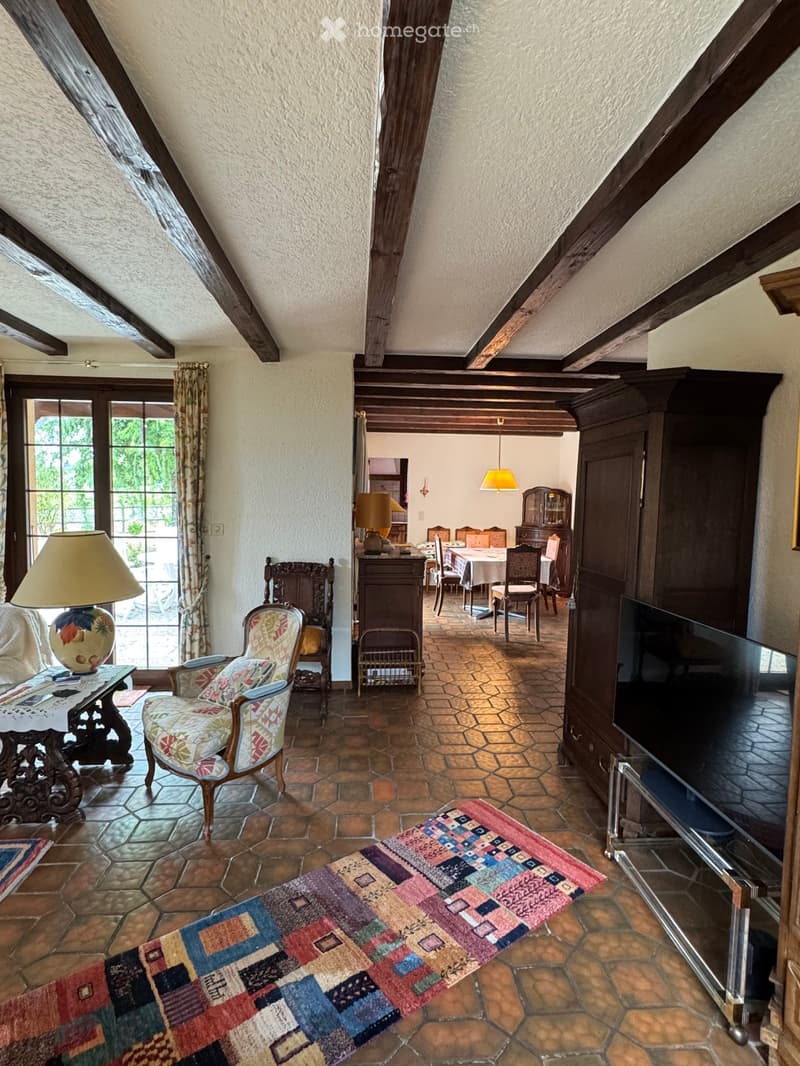 A cozy living room with exposed wooden beams, a patterned rug, a sofa, a coffee table, a television, and a dining area visible in the background.