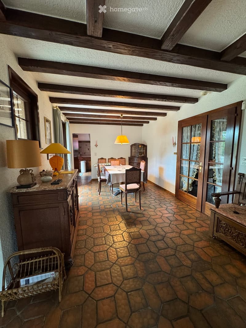 A cozy, rustic dining area with exposed wooden beams, a hexagonal tiled floor, and a dining table set with chairs. A sideboard with a lamp and decorative items is visible, along with a wicker chair and a cabinet with glass doors.