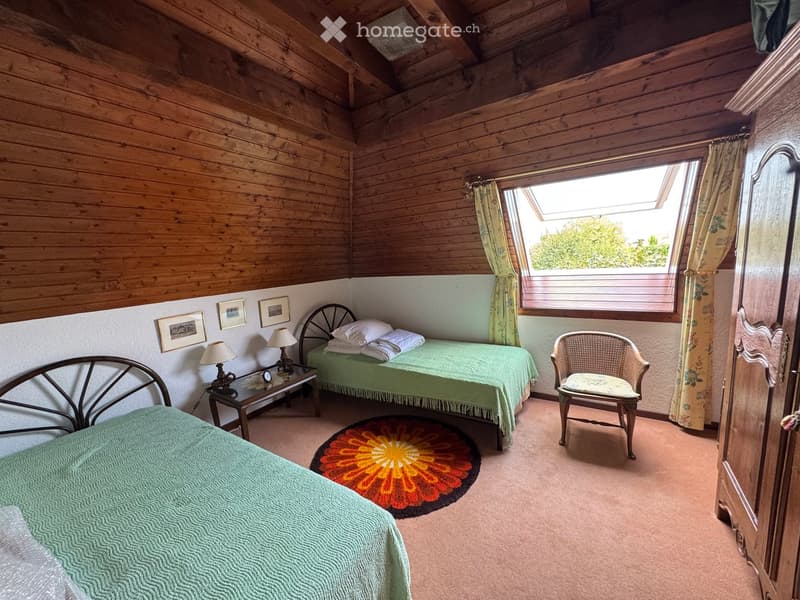 Cozy bedroom with wooden walls and ceiling, two single beds, a wicker chair, a small table with lamps, and a colorful round rug.