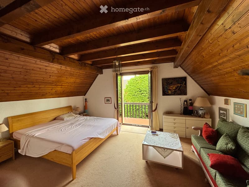 Cozy attic bedroom with wooden beams, a large bed, a sofa, a small table, and a balcony overlooking greenery.