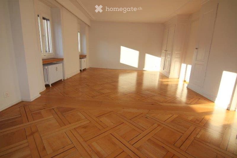 Completely empty room with wooden parquet flooring, white walls, and large windows allowing natural light to fill the space.