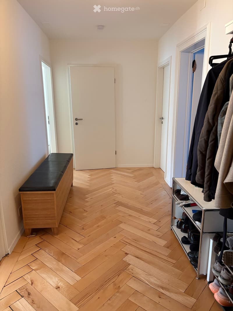 Hallway with herringbone wooden flooring, white walls, and several doors. Features a wooden bench with a black leather seat and a shoe rack with multiple pairs of shoes and jackets.