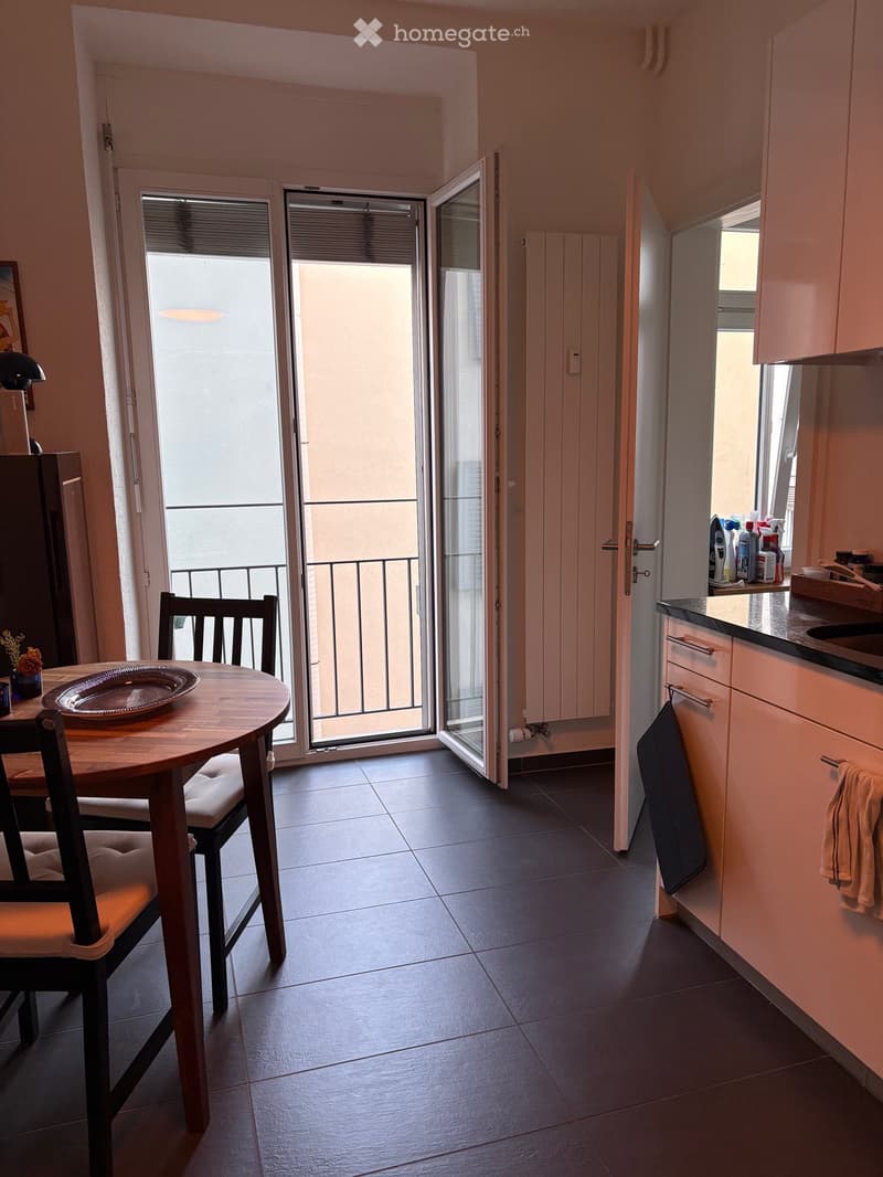 A modern kitchen and dining area with a round wooden table, chairs, and a balcony access. The kitchen features white cabinets, a sink, and various cleaning supplies on the counter.