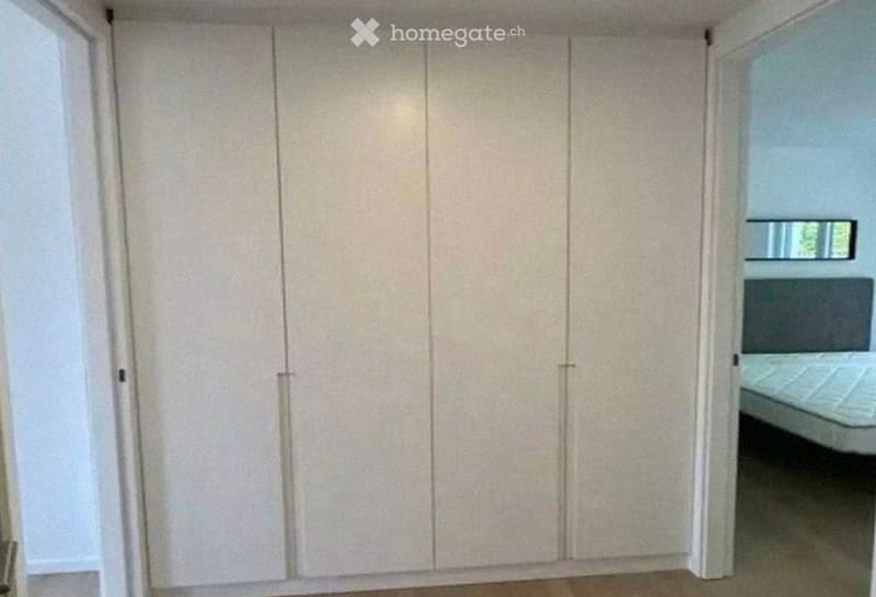 A furnished room with a large, light-colored wardrobe and a bed visible in the background.