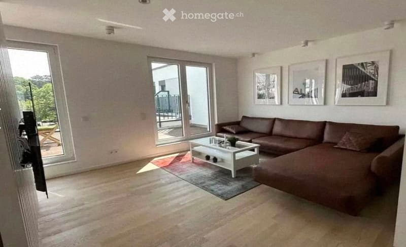Modern living room with large windows and a balcony, featuring a brown sectional sofa, white coffee table, and artwork on the walls.
