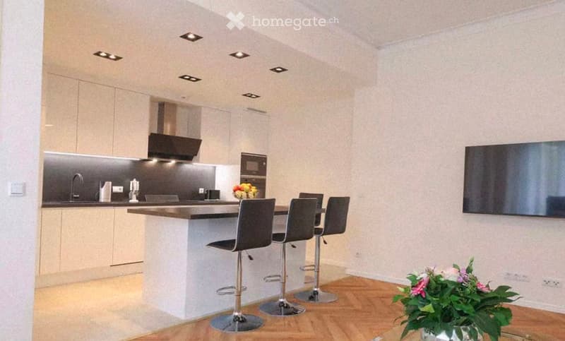 Modern kitchen with white cabinets, stainless steel appliances, island with bar stools, and a flat-screen TV on the wall.