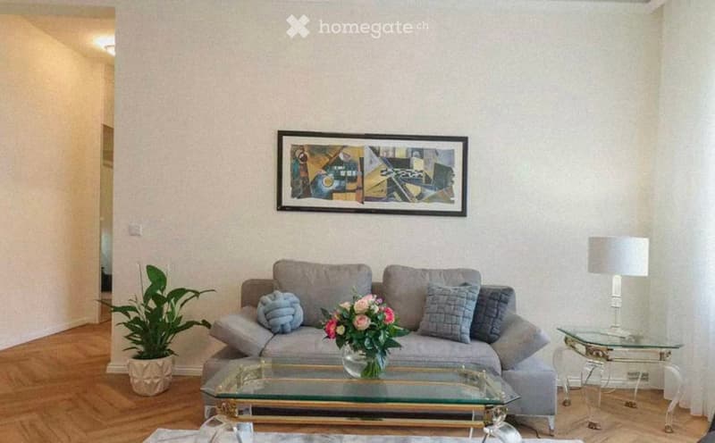 Living room with a grey sectional sofa, glass coffee table, side table with a lamp, potted plant, and a decorative flower vase on the coffee table. A framed abstract artwork hangs on the wall.