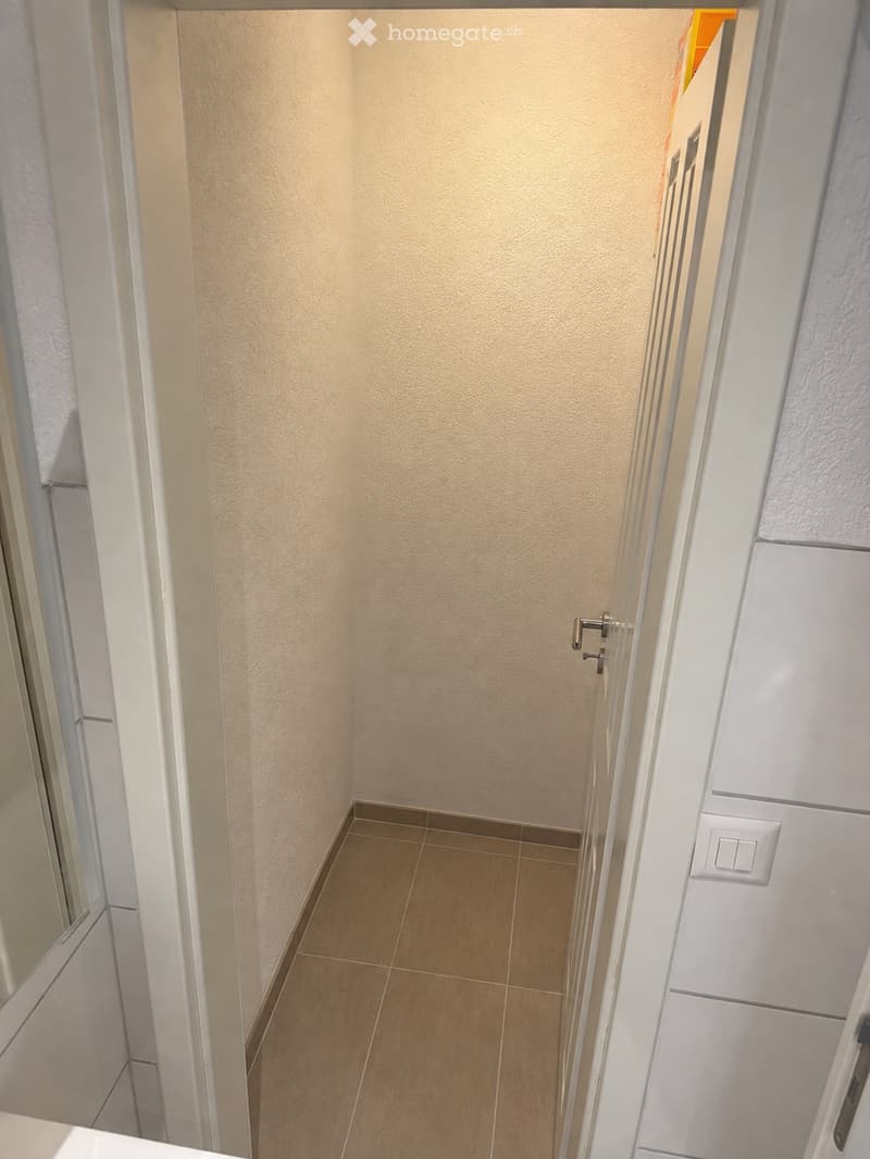 A narrow hallway with beige walls, tiled floor, and white door frames. A light switch is visible on the right wall.