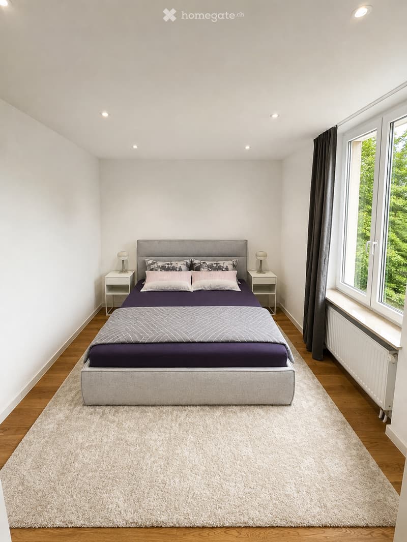 Modern bedroom with a large bed, nightstands, lamps, and pillows, featuring wooden flooring and large windows with curtains.