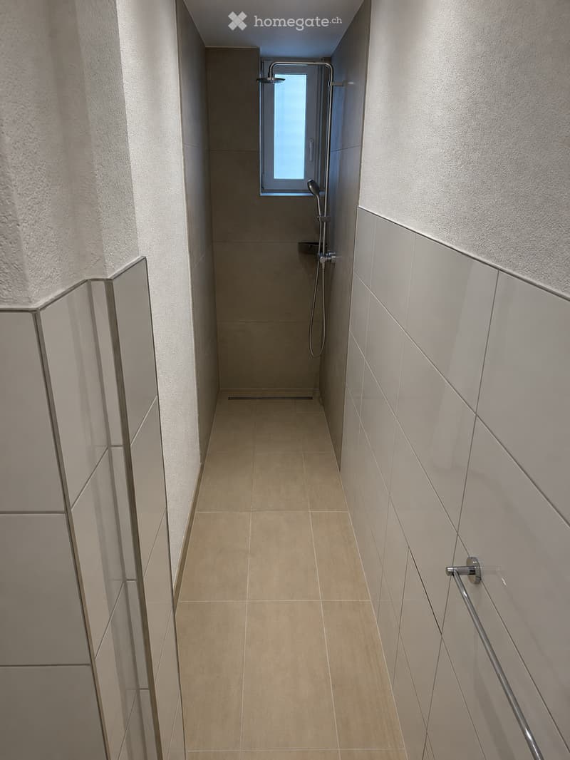 Modern hallway with beige tiled floor, light grey textured walls, and a small window at the end. Features a showerhead and towel rack on the right side.