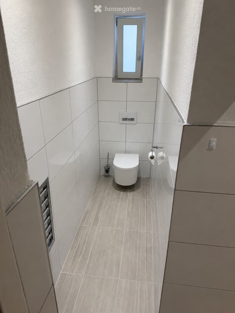 Modern, compact bathroom with white tiled walls, light wood flooring, a toilet, toilet paper holder, and a toilet brush holder.