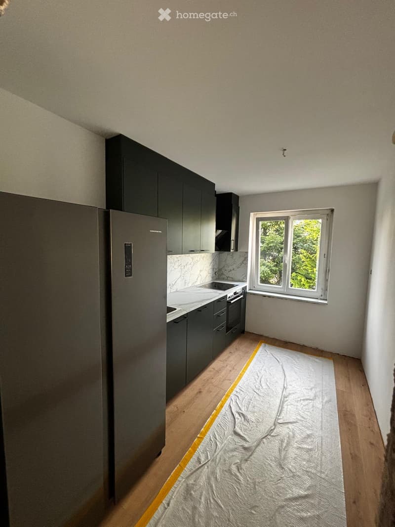 Modern kitchen with dark grey cabinets, a stainless steel refrigerator, and a marble countertop. The floor is wooden, and a protective sheet covers part of the floor.