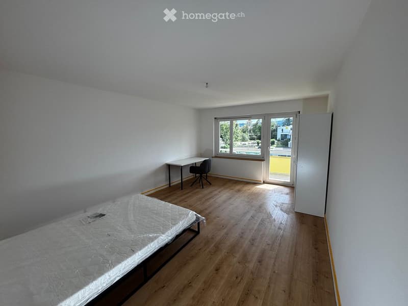 A furnished room with a bed, a chair, and a table, featuring wooden flooring and large windows with a view of the outside.