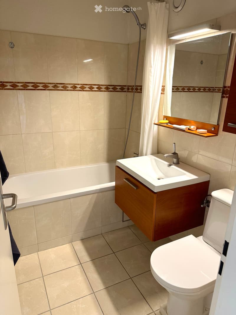 Modern bathroom with beige tiles, a bathtub, a sink with a wooden cabinet, a toilet, a mirror, and a showerhead.