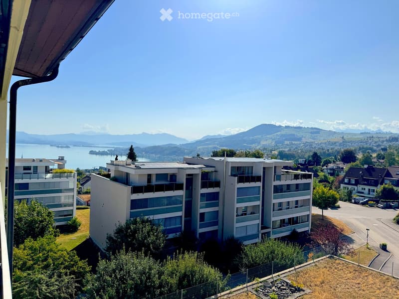 Modern multi-story residential building with balconies and solar panels on the roof, surrounded by greenery and overlooking a lake and mountains.
